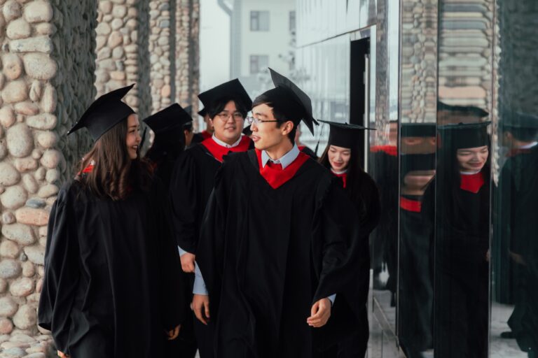 Happy group of graduate students in gowns and mortarboards walking along university corridor and talking
