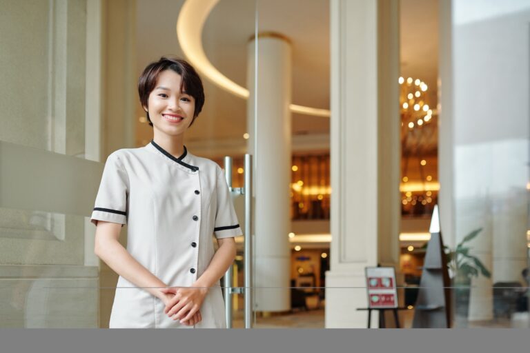 Portrait of cheerful young Asian hotel manager standing at entrance doors and smiling at camera