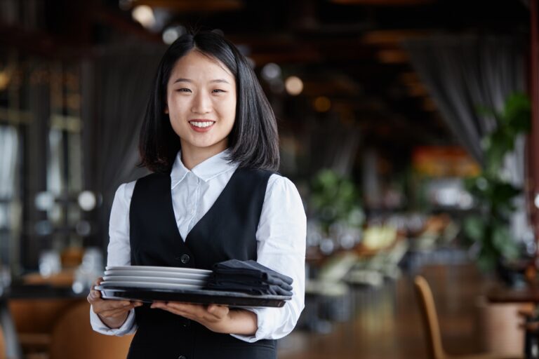 Waist up portrait of young Asian woman as classic server holding tray with plates and smiling at camera in luxury restaurant, copy space