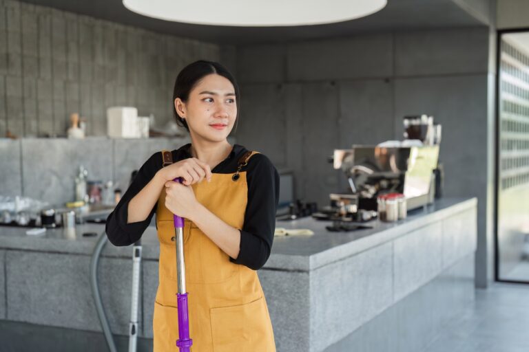 A young barista in a yellow apron cleans a modern coffee shop before opening, ensuring everything is ready for the day.